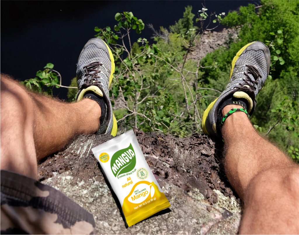 MANGUA a freeze-dried mango on a cliffside with hiker’s legs overlooking a forested valley.